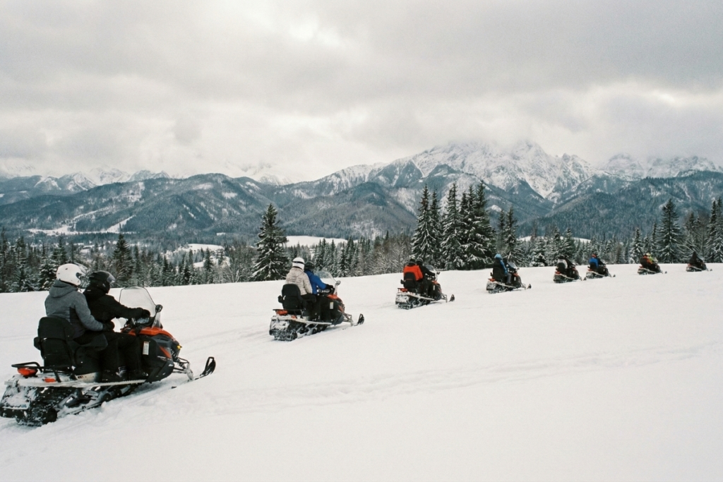 Group of snowmobilers in convoy riding across snowy mountain meadow with Tatra Mountains landscape in background